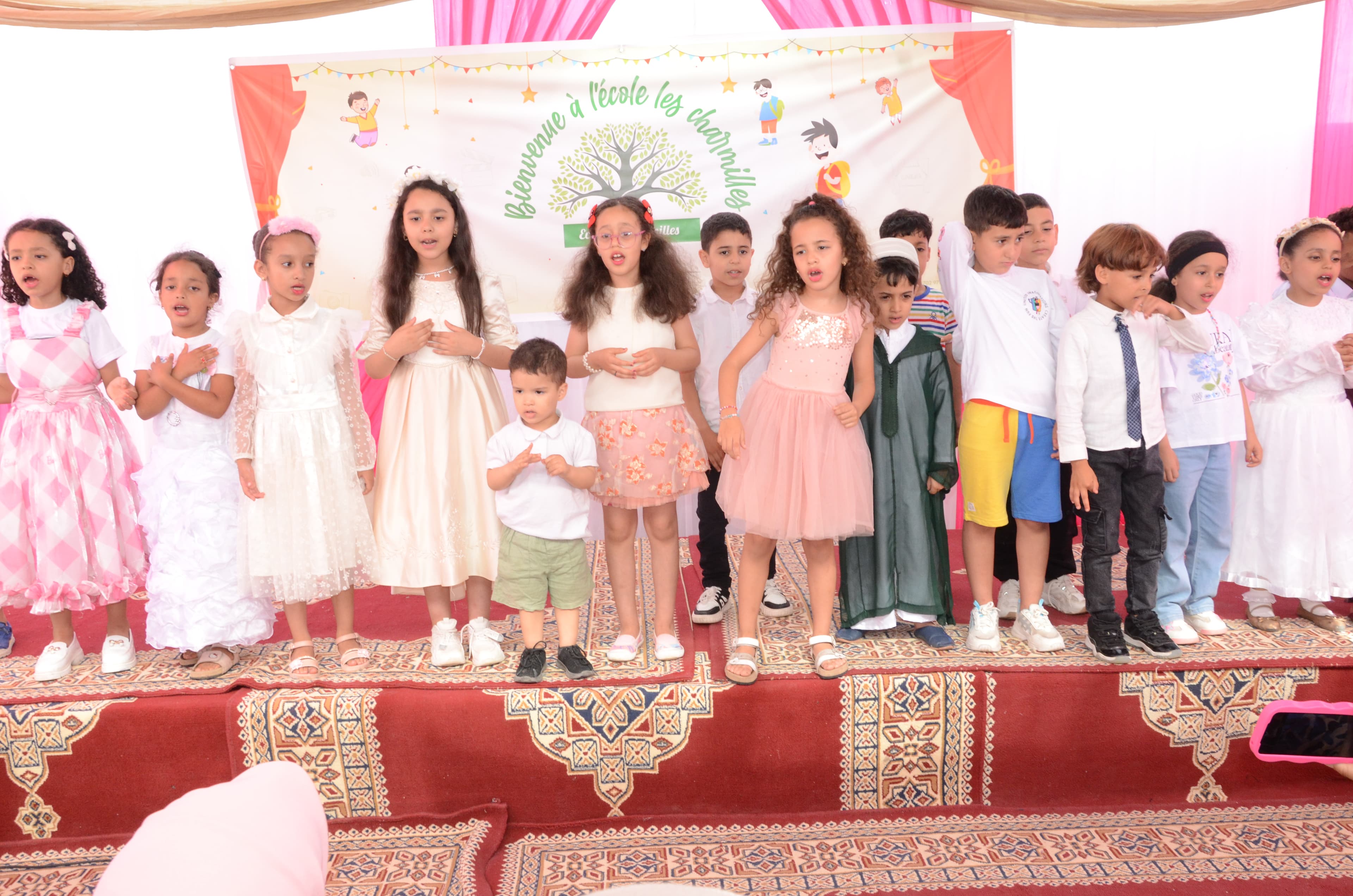 Grand groupe d'enfants en tenues de fête variées, chantant ou posant ensemble sur la scène lors d'un événement scolaire.